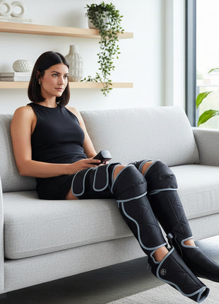 Woman using an electric leg massager on a dark gray couch in a living room setting.